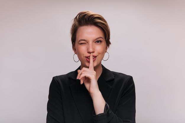 Woman winking and asking for keeping secret. Short-haired lady in stylish jacket posing and looking into camera on isolated