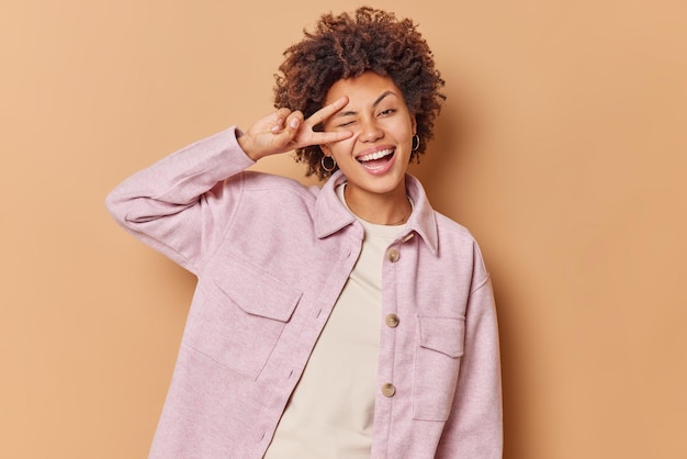 woman winks eye makes peace sign smiles broadly shows white teeth wears pink jacket enjoys life expresses positive emotions isolated on beige shows salute gesture