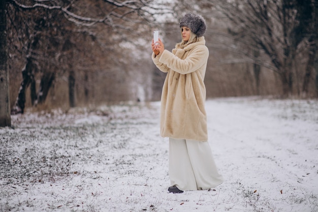 電話で話している雪でいっぱいの公園を歩く冬のコートの女性