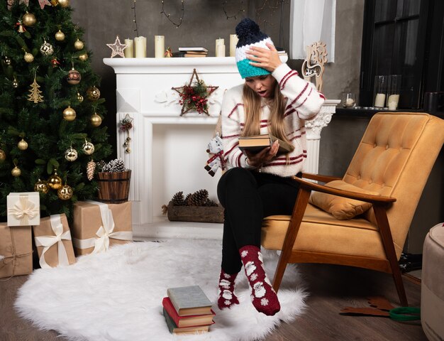 Woman in winter outfit checking names of books near the fireplace. 