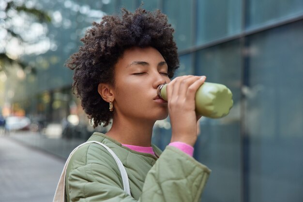 woman with Afro hair drinks water from bottle takes break while walking in city poses in urban setting keeps eyes closed wears jacket hydrates herself