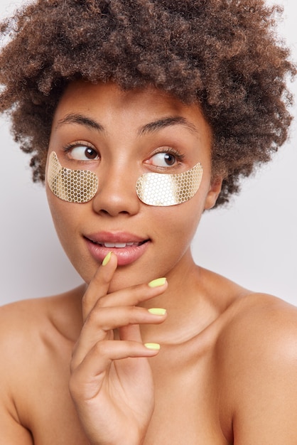 woman with Afro hair keeps hand on chin looks away pensively undergoes beauty procedures applies patches under eyes to moisturize skin stands topless indoor