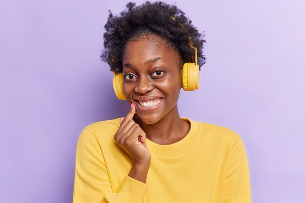  woman with Afro hair smiles gladfully shows white teeth listens audio track in wireless headphones wears casual jumper isolated on purple 