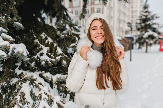 に覆われた木がある公園で冬の休暇を過ごす素晴らしい笑顔を持つ若い女性。寒い日に新鮮な空気を楽しんでいる長い髪の喜んでいるヨーロッパの女性の屋外のポートレート。