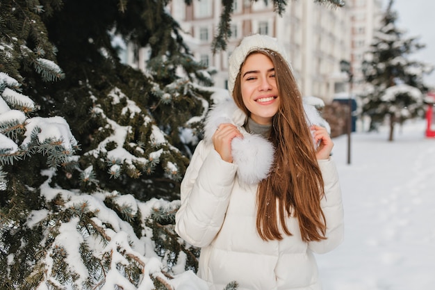 woman with amazing smile spending winter vacation in park with snowy trees. Outdoor portrait of glad european woman with long hair enjoying fresh air in cold day.