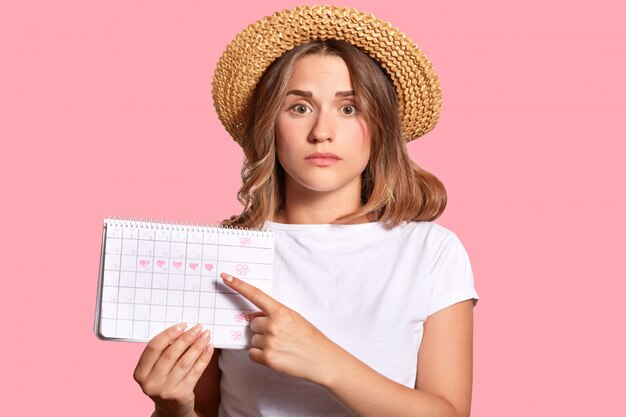 Woman with appealing look, holds periods calendar for checking menstruation days, points with fore finger