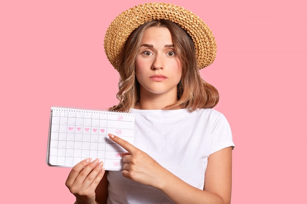 Woman with appealing look, holds periods calendar for checking menstruation days, points with fore finger