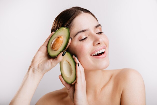 Женщина с голыми плечами мило улыбается, позируя с авокадо, полезным для кожи на белом фоне