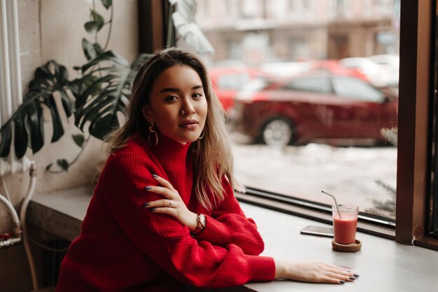 Woman with black manicure dressed in red knitted sweater posing at table in cafe by window