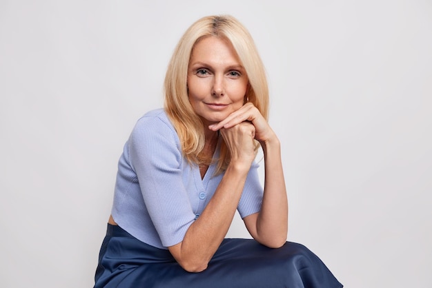 Free photo woman with blonde hair keeps hands near face looks directly at camera wears blue jumper and skirt sits on white thinks about something