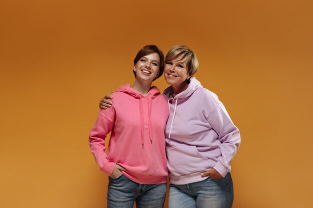 Woman with blonde hair in lilac sweatshirt and jeans smiling and hugging with young girl with brunette hairstyle and pink hoodie.