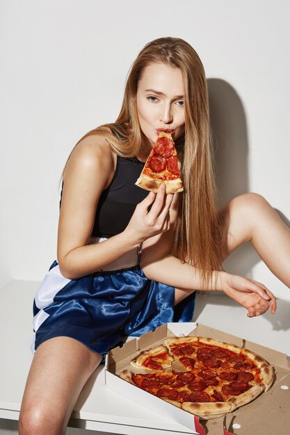 금발 머리 테이블에 확산 다리 앉아, 꼬리 치는 식으로 피자를 먹는 여자.