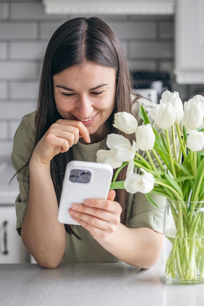 Free photo woman with a bouquet of white tulips uses a smartphone at home in the kitchen