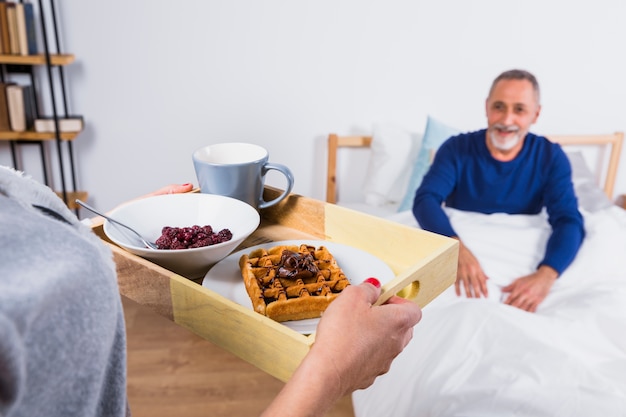 Donna con colazione vicino uomo sorridente invecchiato in piumone sul letto