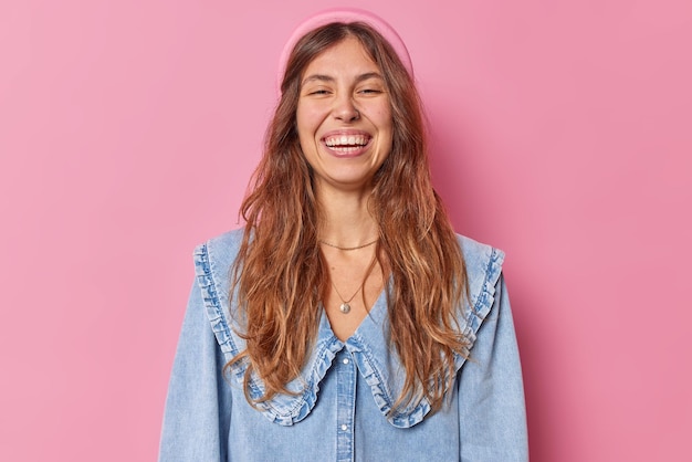 woman with broad smile shows perfect white teeth wears denim shirt and headband isolated on pink has piercing in nose shows happy emotions. Studio shot