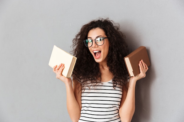 갈색 곱슬 머리는 회색 벽 위에 절연 교육에 즐거움을 복용 손에 재미있는 책과 함께 포즈 대학에서 학생이되는 여자