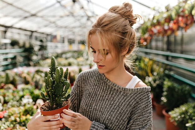 お団子を持つ女性は、植物のあるギャラリーを歩きながら、興味を持って鍋にサボテンを見ています。