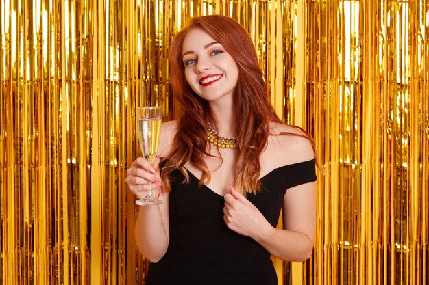 Woman with charming smile celebrating new year, holding glass of wine, wearing elegant black dress, posing against yellow wall with golden tinsel.