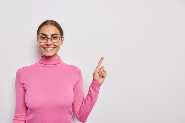 woman with cheerful expression says look here indicates at upper right corner advertises your text poses on white wears pink turtleneck and round spectacles