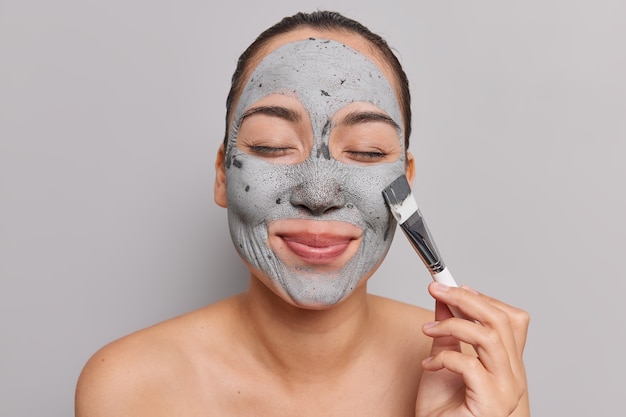 woman with combed hair keeps eyes closed applies clay mask on face holds cosmetic brush takes care of complexion stands shirtless indoor on grey 