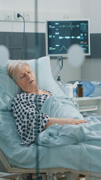 코로나바이러스 증상이 있는 여성이 병원 병동 폐쇄 상태에서 심하게 숨을 쉬면서 의료 도움을 요청합니다. 침대에 누워 과호흡하는 바이러스에 대한 고립된 지역의 노인 환자