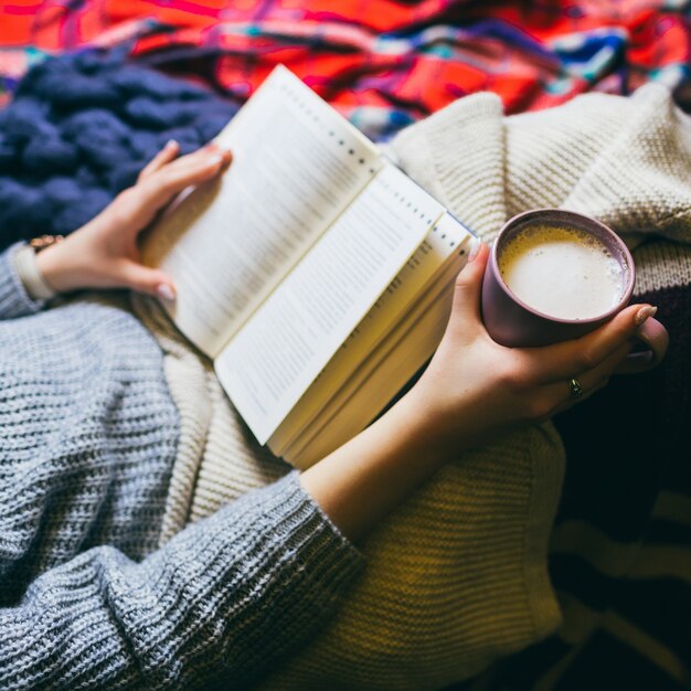 Woman with cup of coffee and book lies under colorful plaids