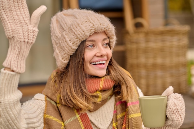 Woman with cup looking aside raising hand in greeting