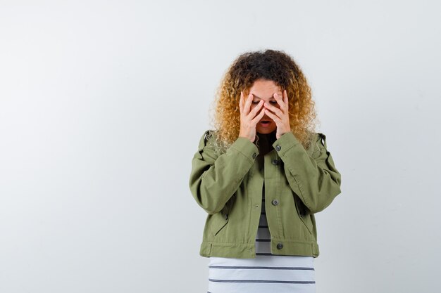 Woman with curly blonde hair in green jacket keeping hands on face and looking upset , front view.