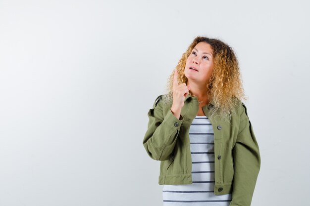 Woman with curly blonde hair in green jacket pointing up and looking wondered , front view.