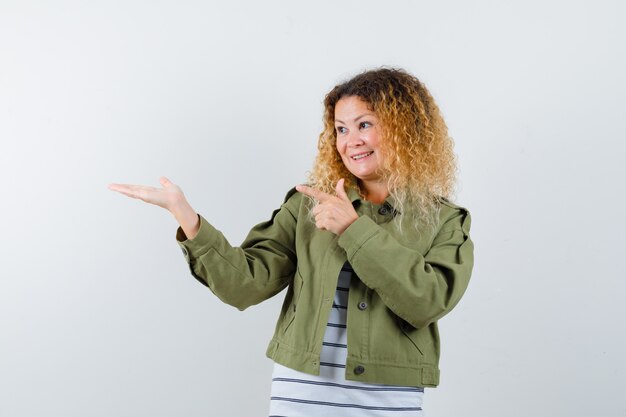 Woman with curly blonde hair pointing at her palm spread aside in green jacket and looking jolly , front view.