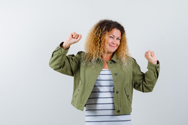 Woman with curly blonde hair showing winner gesture in green jacket and looking merry , front view.