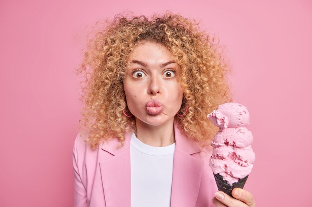 woman with curly bushy hair keeps lips folded stares at camera holds big appetizing cone ice cream in waffle wears formal jacket isolated on pink 