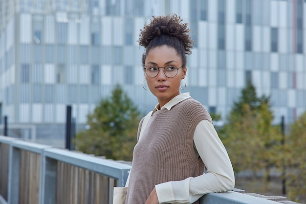 Woman with curly combed hair wears roud spectacles for vision correction wears neat clothes poses on bridge against urban environment