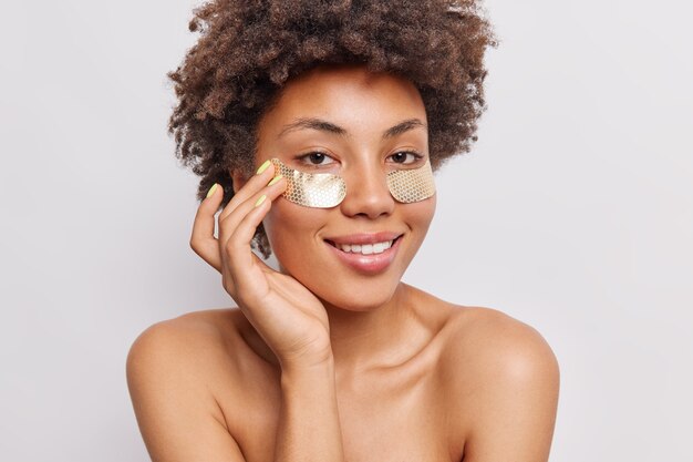 woman with curly hair applies beauty hydrogel patches under eyes smiles gently stands topless on white 