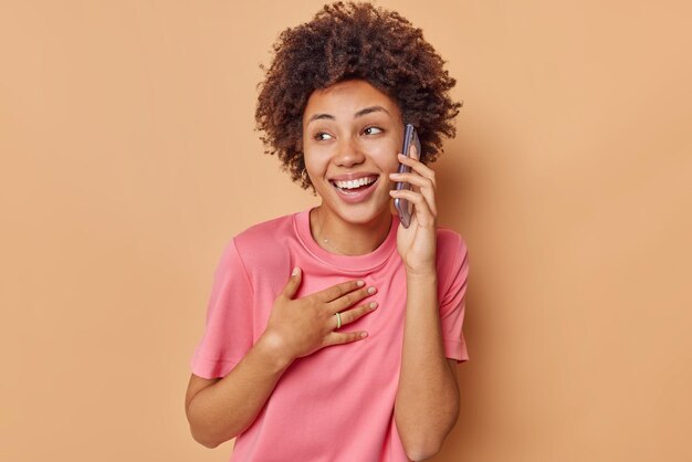 woman with curly hair calls via smartphone discusses something positive with friend wears casual pink t shirt isolated on beige. People and communication concept