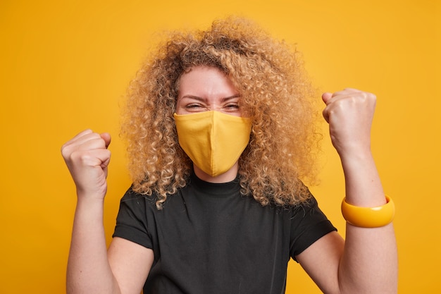 La donna con i capelli ricci stringe i pugni con trionfo celebra il successo indossa una maschera protettiva contro il coronavirus la maglietta nera posa contro lo sfondo giallo. mantieni le misure di quarantena