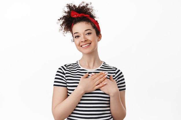 Donna con i capelli ricci pettinati, fascia per capelli, tenendosi per mano sul cuore e guardando grata, ringraziandoti, essendo toccata o lusingata, in piedi sul bianco