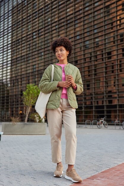 woman with curly hair dressed in stylish apparel holds paper disposable cup of coffee looks away carries fabric bag stands on pavement in city walks outside