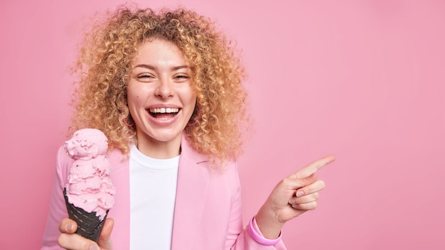 woman with curly hair eats delicious ice cream in waffle points away on blank space presents summer product wears pink jacket 