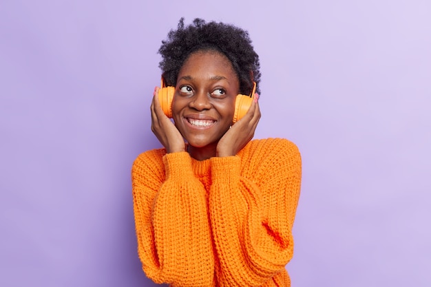 Woman with curly hair enjoys listening audio track keeps hands on headphones thinks about something good wears knitted orange sweater isolated on purple