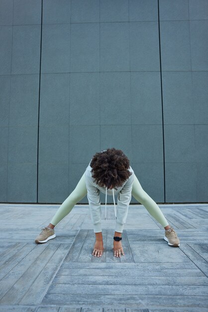 都会の灰色の壁に対して巻き毛のエクササイズをしている女性は、スポーツウェアを着た床に寄りかかって、屋外でジョギングする前にアクティブなライフスタイルが筋肉を温めることを示しています