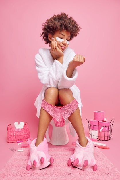 Бесплатное фото Женщина с кудрявыми волосами с расстройством пищеварения сидит на унитазе в туалете, накладывает патчи под глаза, носит белый халат, тапочки, трусики на ногах, изолированные на розовой стене