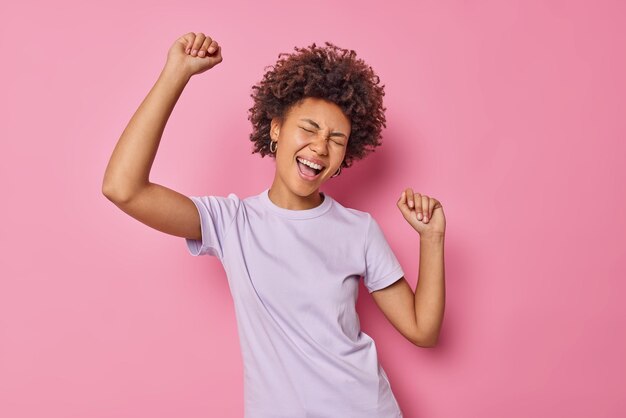 woman with curly hair has fun and dances carefree dressed casually isolated on pink enjoys party and favorite music isolated on pink. People and emotions