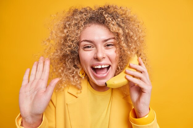 woman with curly hair has happy mood waves palm in hello gesture greets someone holds banana as if telephone 