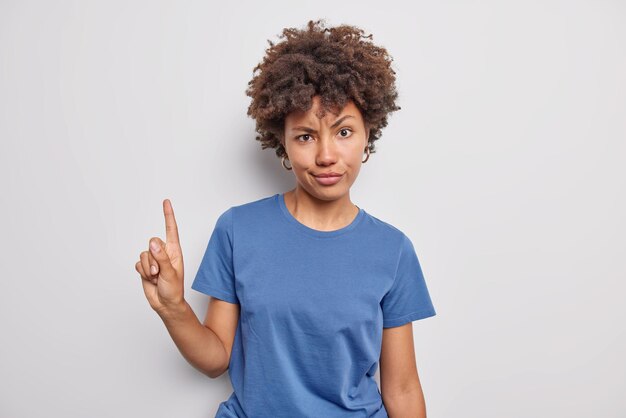 곱슬 머리를 가진 여자는 검지 손가락으로 위의 음침한 표정을 나타냅니다. 흰색으로 격리된 캐주얼한 파란색 티셔츠를 착용합니다.