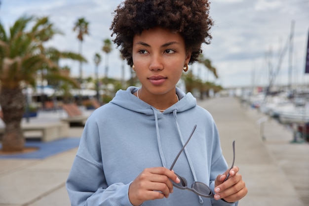 woman with curly hair holds sunglasses dressed in hoodie walks near sea port during daylight spends weekends actively