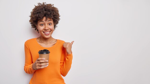 женщина с вьющимися волосами указывает большой палец на пустую копию космоса пьет ароматный кофе на вынос носит оранжевый джемпер, изолированный на белом. Посмотри на это