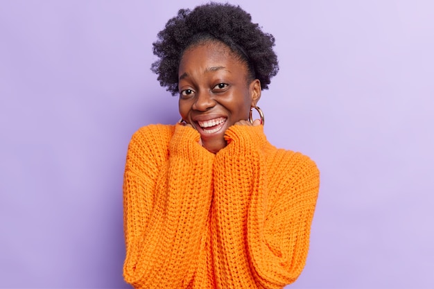 La donna con i capelli ricci tiene le mani sotto il mento sorride mostra positivamente i denti bianchi ha buon umore indossa un maglione lavorato a maglia arancione di grandi dimensioni isolato su viola