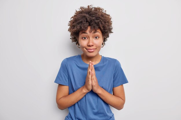 woman with curly hair keeps palms pressed together thanks someone believes in good news wears casual blue t shirt isolated on white. Please give me one more chance