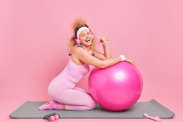 Woman with curly hair leans on fitness ball being in good mood listens music via wireless headphones exercises at mat